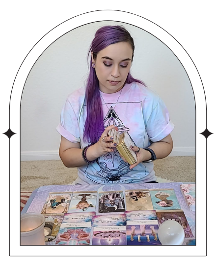 Ellie sits at a small table, holding tarot cards mid-shuffle. The image captures a moment of focused intention, with warm lighting that highlights the reflective and compassionate approach to tarot readings offered by Ellie.